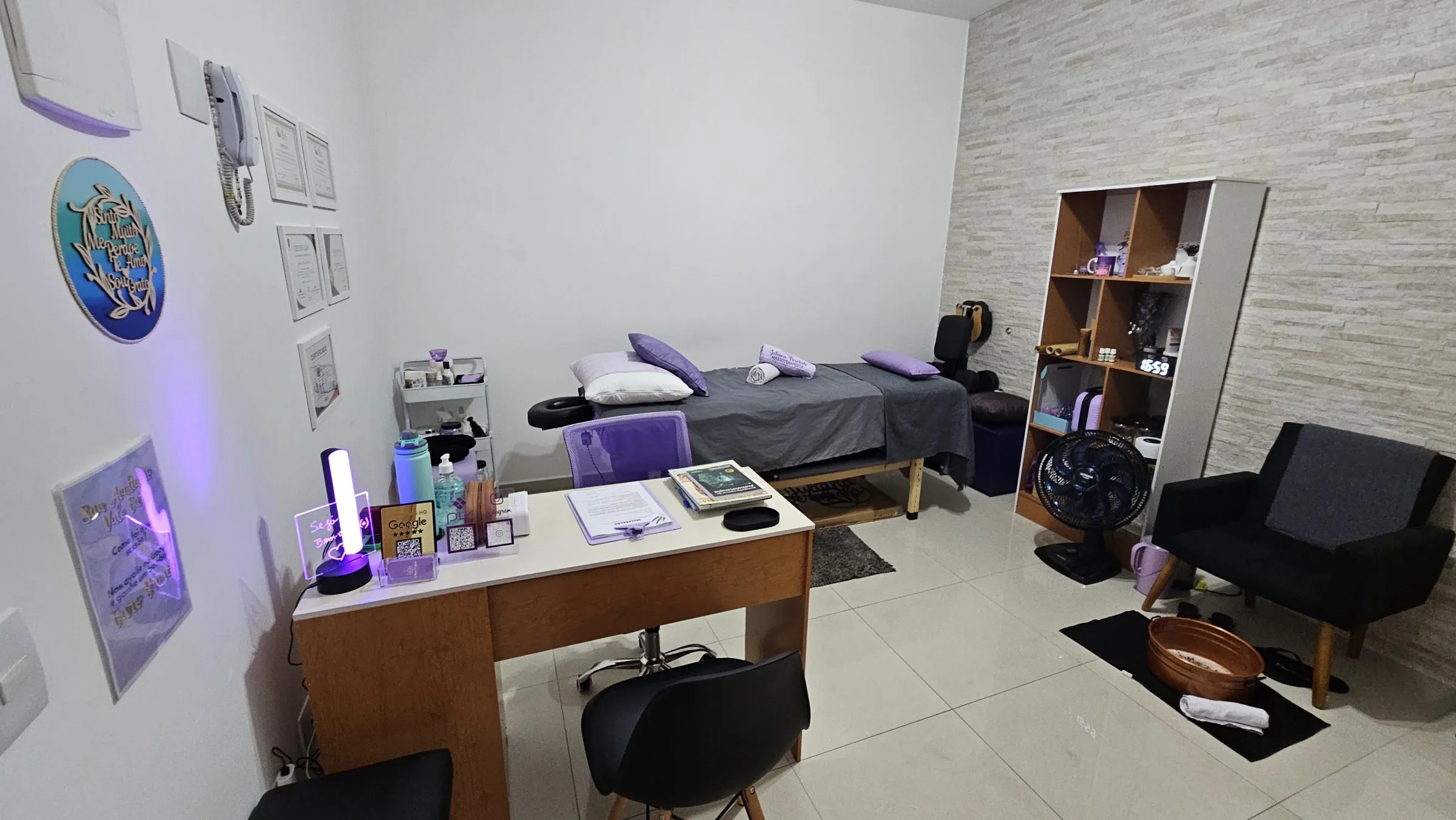 Sala de atendimento do Espaço Juliana Perobelo: mesa de conversa à esquerda, maca com almofadas lilás ao fundo, poltrona e bacia de cobre para escalda-pés à direita